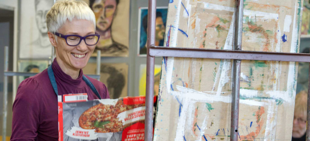 Kuopion kansalaisopisto, kuvataide. Hymyilevä nainen maalaustelineen edessä.